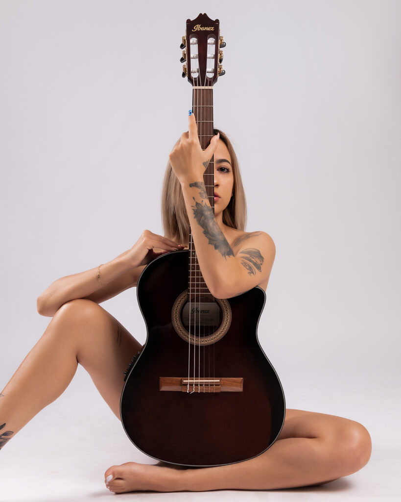 Woman sitting with a classical guitar, partially obscuring her face, showcasing tattoos and elegance, photographed at Duette Studio in Miami.