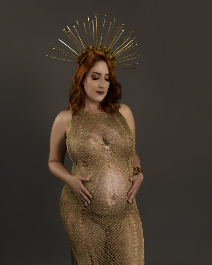 A pregnant woman in a golden gown and a celestial crown cradles her baby bump, standing against a dark background. A majestic maternity session by Duette Studio.
