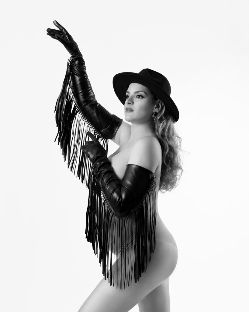 A pregnant woman in a black hat and leather fringe gloves poses dramatically in a black-and-white artistic maternity shoot. A bold and unique maternity session by Duette Studio.