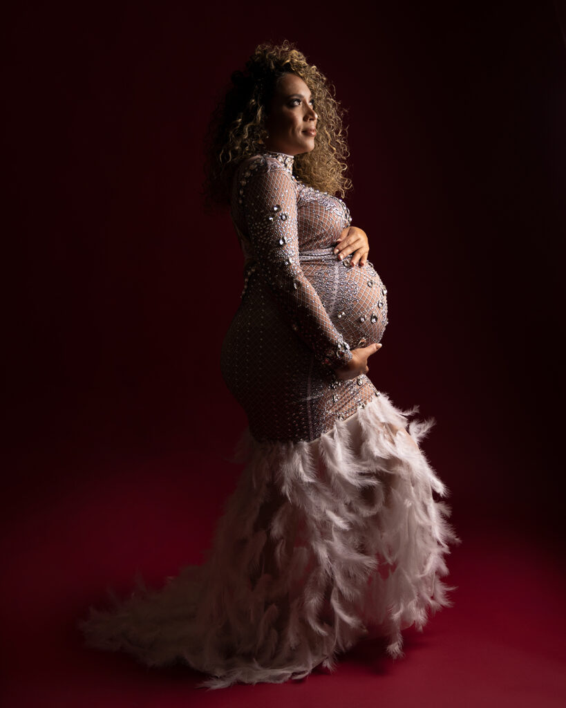 A pregnant woman in a shimmering, feathered gown cradles her baby bump against a deep red background, looking serene and radiant. A glamorous maternity session by Duette Studio.